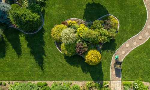 Garden Maintenance and Care Work in Process. Wheelbarrow with Grass Cuttings and a Plants Watering Can Stand on a Twisting Garden Path Next to a Large Landscape Island. Aerial View.